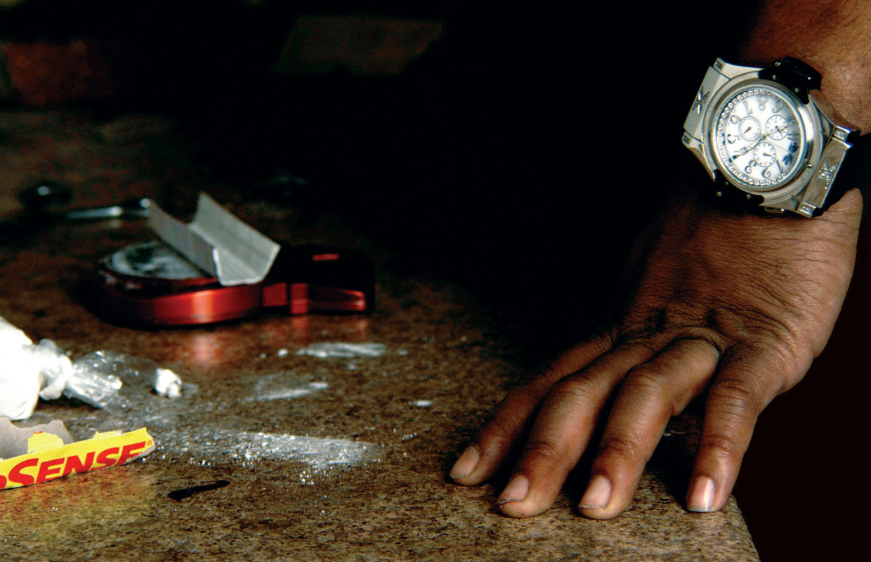 A hand on a counter next to a grinder, rolling papers, and a white powder. A scene from the movie How To Make Money Selling Drugs