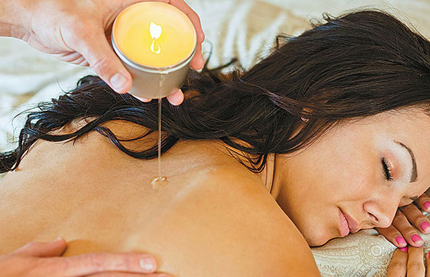A dark haired woman lies face down on a massage table has hot wax poured on her bare back from Earthly Bodies 3-in1 Candle