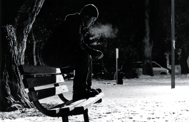 A black and white night-time image of a man sitting on the back of a park bench.