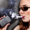 A brunette woman with sunglasses and red lips blows smoke from a Da Vinci Vaporizer