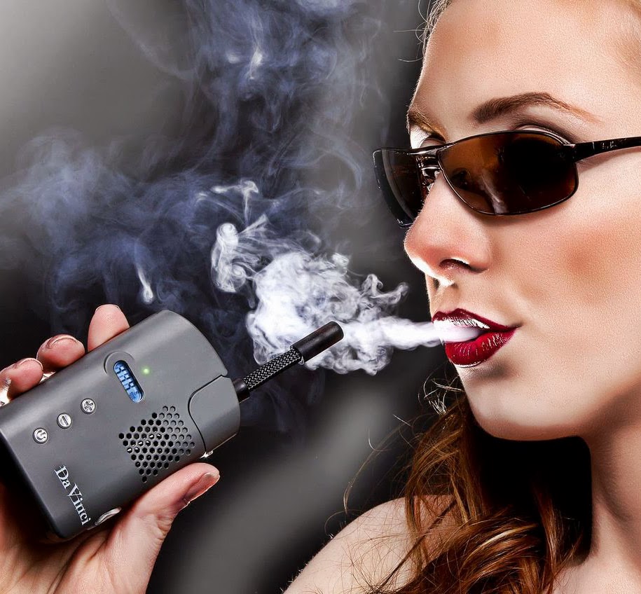A brunette woman with sunglasses and red lips blows smoke from a Da Vinci Vaporizer