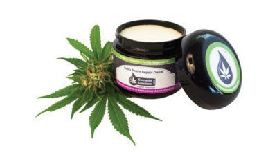 A pot of body butter by Cannabis Creations sits on top of and next to a pot leaf and flower.
