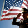 An veteran dressed in uniform salutes while standing in front of a flag.