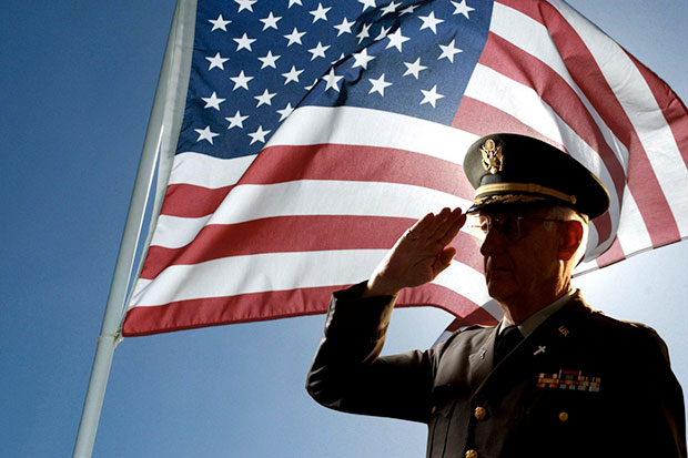 An veteran dressed in uniform salutes while standing in front of a flag.