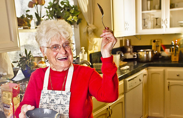 Nonna Marijuana Wears a Red Shirt and Apron While Smiling and Holding Up a Tasting Spoon