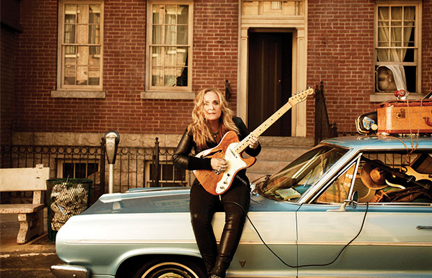 Melissa Etheridge Sits Atop Her Car Playing A Guitar in Front of a Brick Building