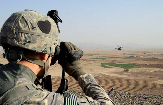 A soldier uses binoculars to look out over a vast landscape at a helicopter in the distance. Perhaps he is also looking to a future where veterans have access to MMJ.