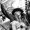 A black and white image of Willie Nelson singing on stage with his guitar.