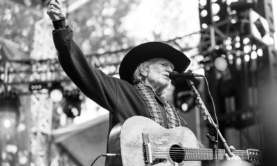 A black and white image of Willie Nelson singing on stage with his guitar.