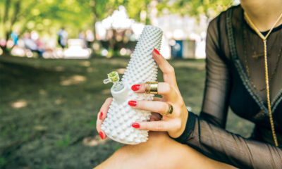 A woman sits on a park bench on a clear day holding a textured and white bong made from a 3-D printer.