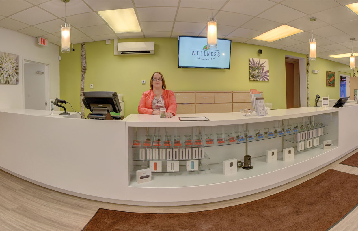 A woman in pink sits behind a white front counter at the Wellness Connection ready to welcome patients.