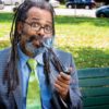 Ngaio Bealum puffs on a pipe while wearing a blue suit and green tie on a park bench as he answers Dear Dabby questions.