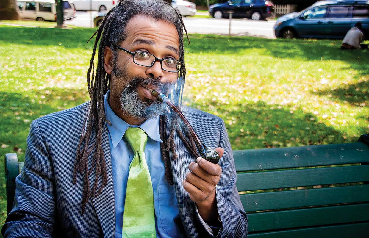 Ngaio Bealum puffs on a pipe while wearing a blue suit and green tie on a park bench as he answers Dear Dabby questions.